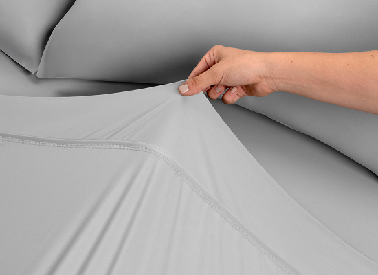 A close-up of a hand stretching a light gray fitted sheet over gray pillows to demonstrate the fabric's elasticity and snug fit.