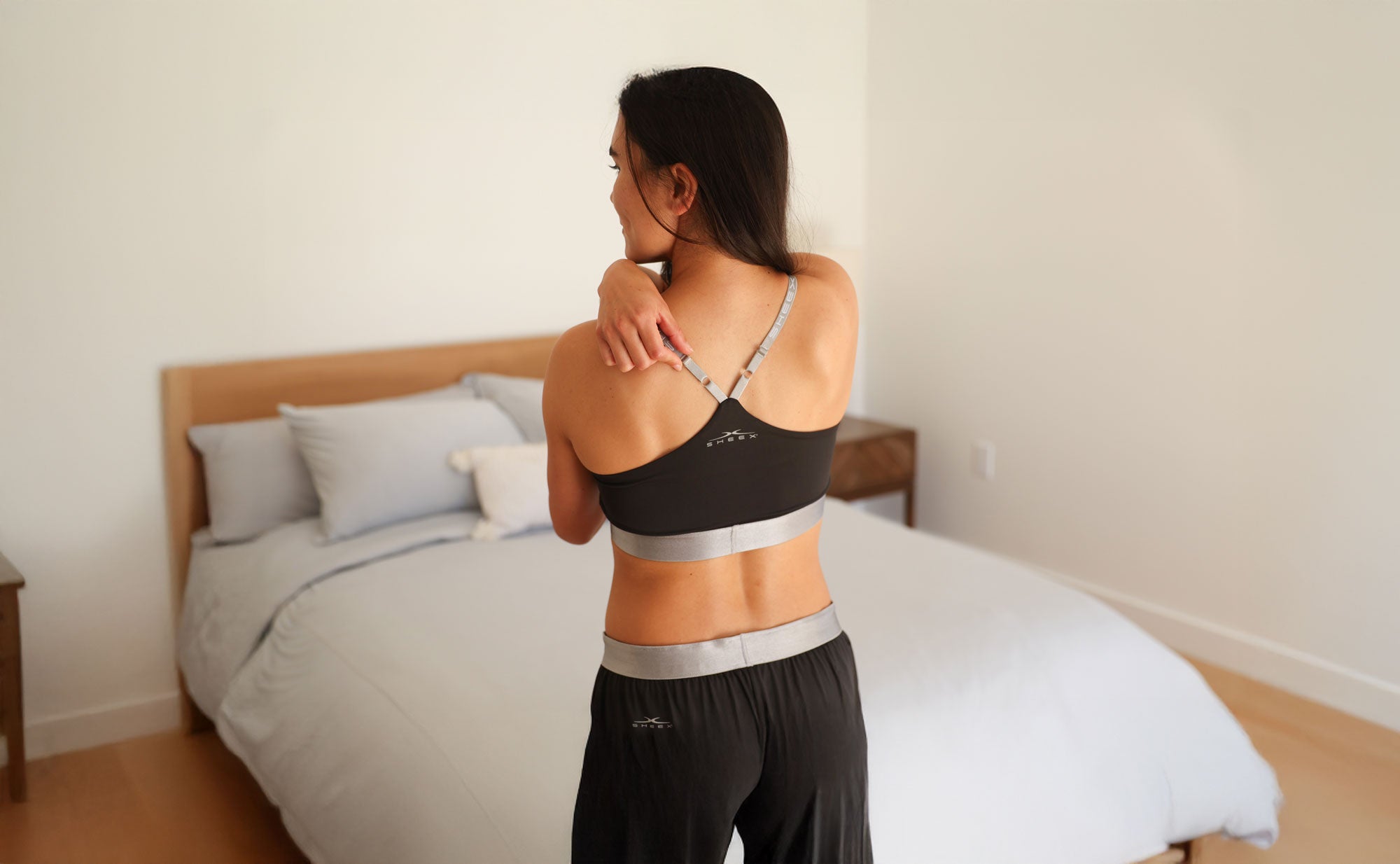 Female standing in bedroom in SHEEX Bra Top and Jogger. Adjusting strap on bra.