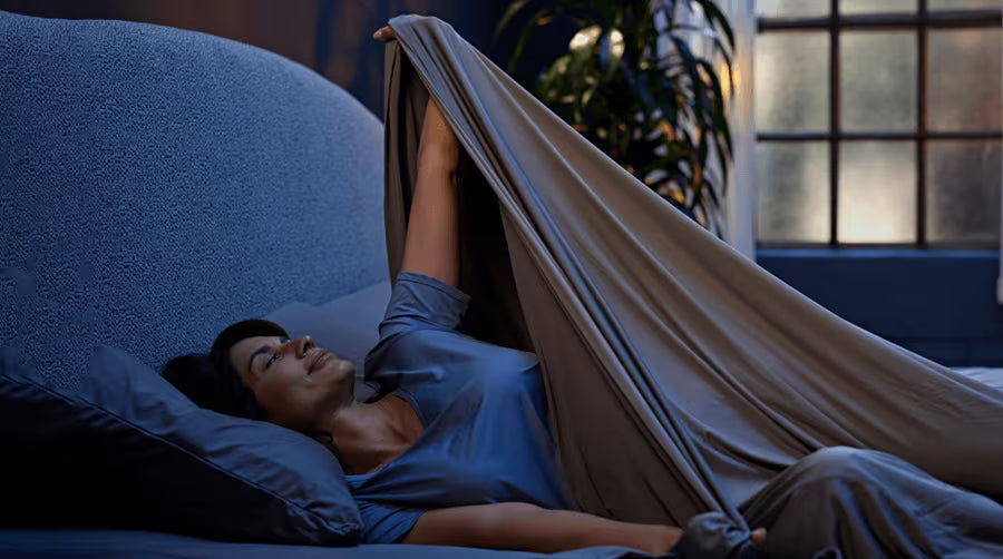 Model lying in bed with Original Performance sheets pulled up to their chest, in a dimly lit room.
