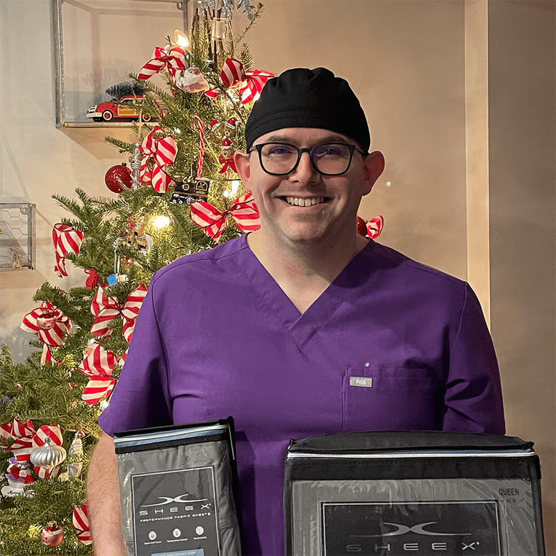 Person in purple scrubs holding SHEEX products in front of a decorated Christmas tree.